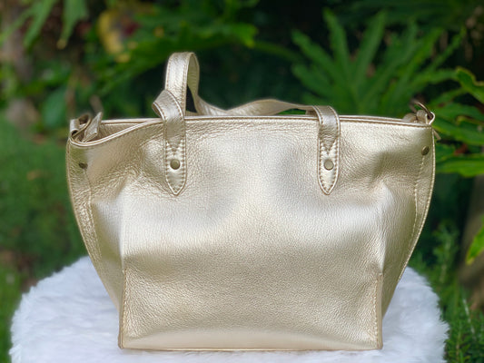 Pale gold metallic leather tote bag on a white surface with a green leafy background