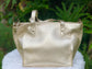 Pale gold metallic leather tote bag on a white surface with a green leafy background