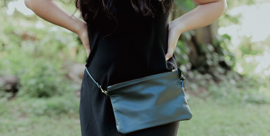Close-up of a woman in a black dress wearing a sleek black leather pouch as a belt bag around her waist, standing in a lush green outdoor setting.