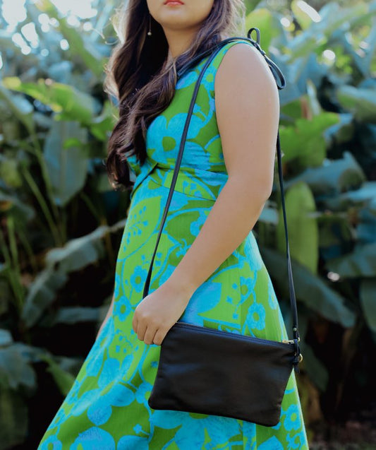 Woman wearing a blue and green patterned dress holding a black pouch on her shoulder with a blurred green plant background