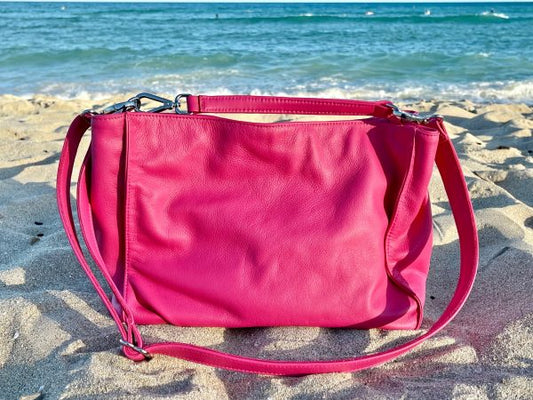 Front view of the hibiscus pink bag   on a sandy beach with ocean in the background. featuring both long crossbody strap and short shoulder strap.